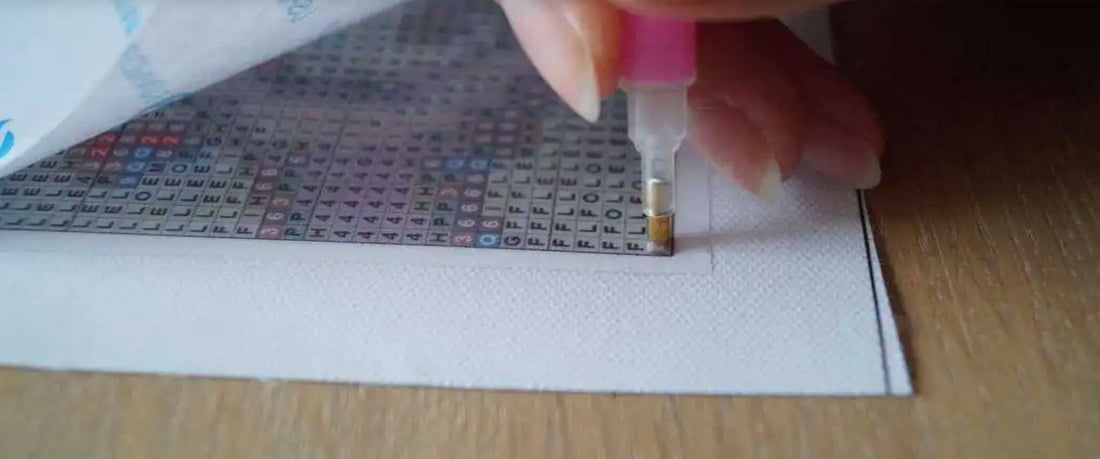 Close-up van een diamant schilderij in progressie, met een hand die een roze pen gebruikt om gekleurde resin diamanten op een genummerd canvas te plaatsen.